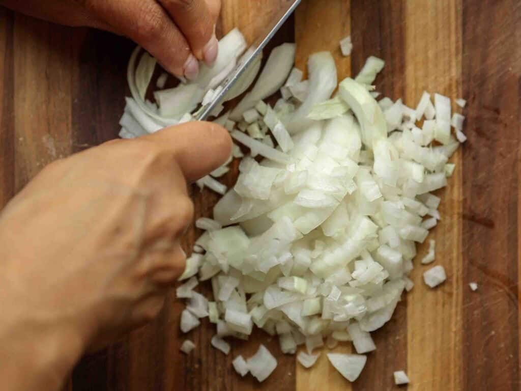 A person’s hands are chopping a white onion into small pieces on a wooden cutting board, prepping ingredients for a vegan bolognese. Some onion slices and diced pieces are visible.